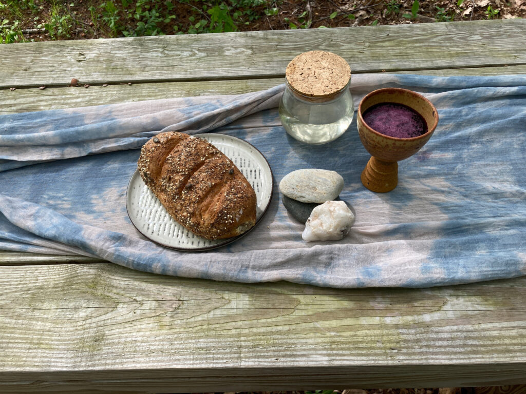 Bread, a chalice and a jar set on a cloth on a wooden picnic table outdoors