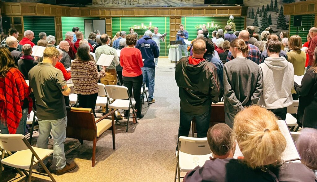 A full room of worshipers standing and singing at PEACE Mennonite Fellowship